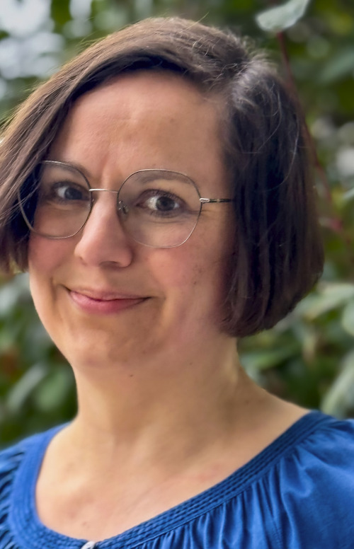 Ein Bild von mir. Ich habe mittellange braune Haare, die auf der Seite gescheitelt sind. Ich trage eine Brille mir schmalem Metalrand und ein blaues Oberteil.