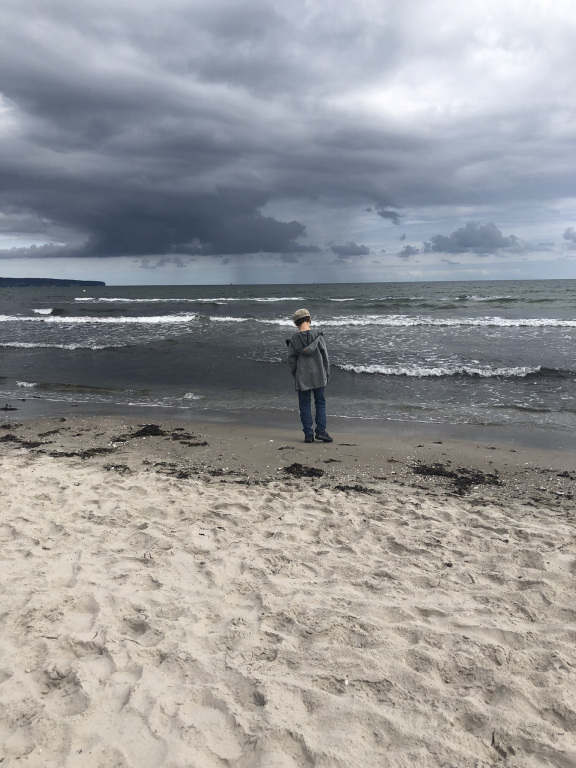 unser Kind von hinten an einem stürmischen Tag am Ostseestrand, der Blick geht aufs Wasser, dunkle, tiefhängende Wolken